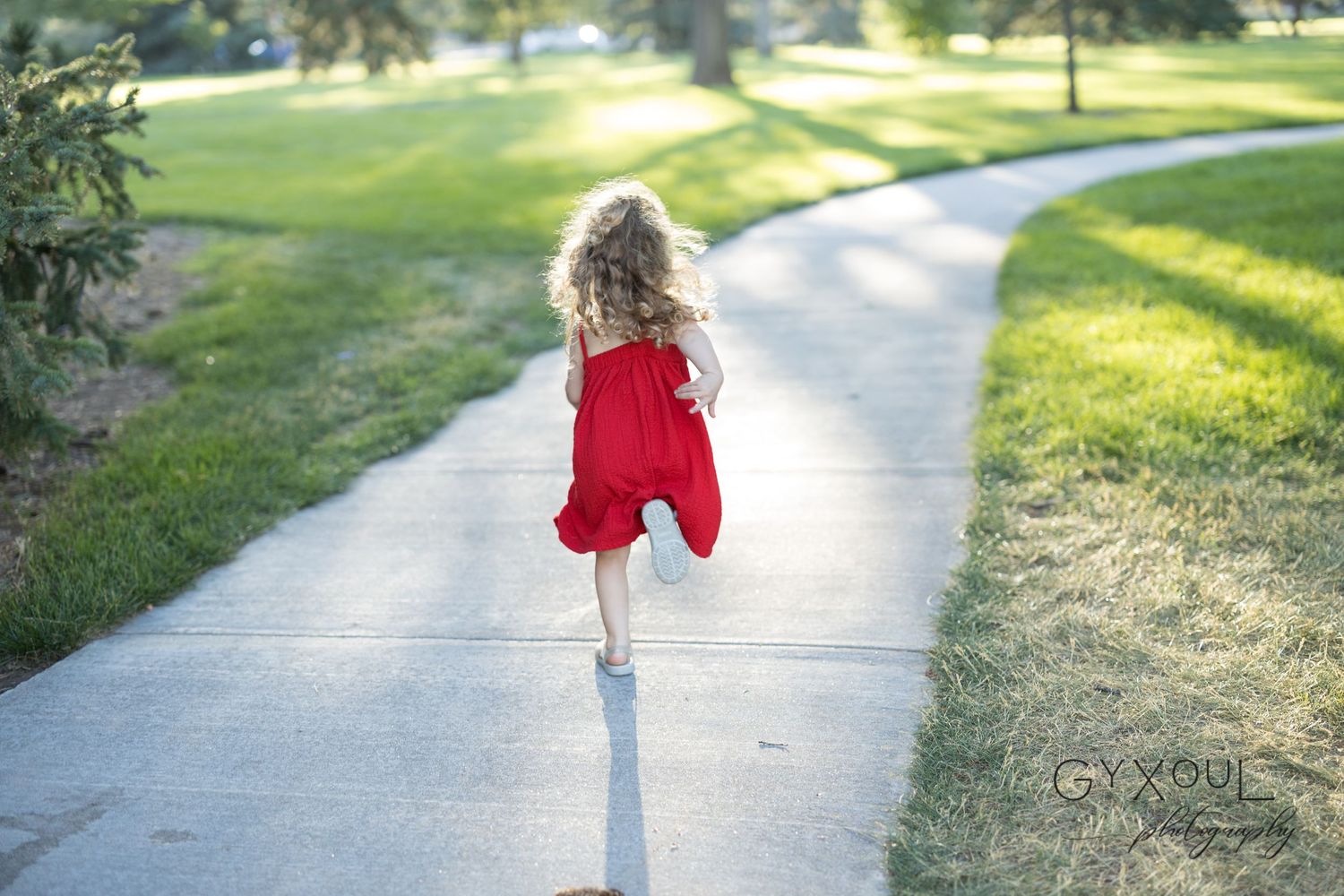 A child in a flowing red dress runs along a sun-lit walking path.