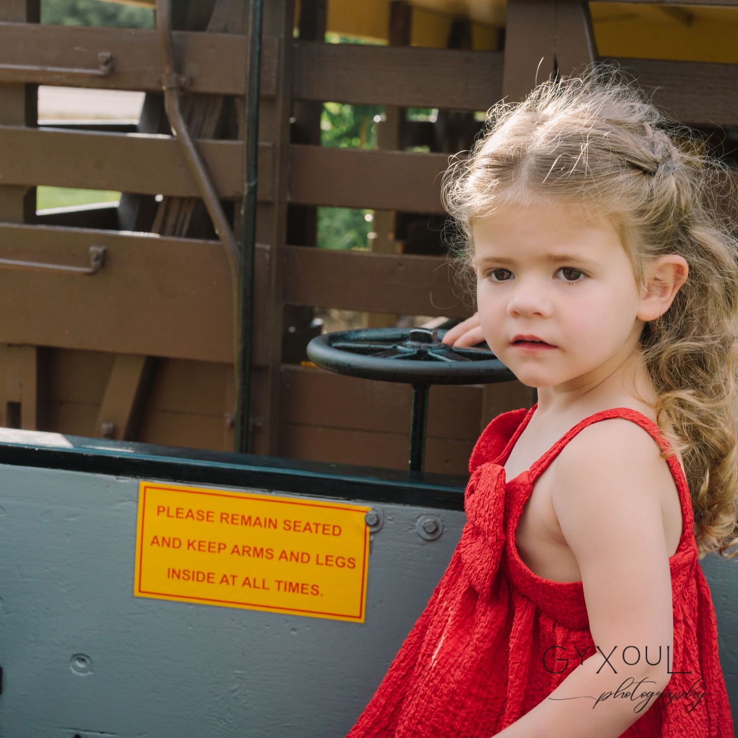 A child in a red sundress stands near a wooden structure with a yellow warning sign.