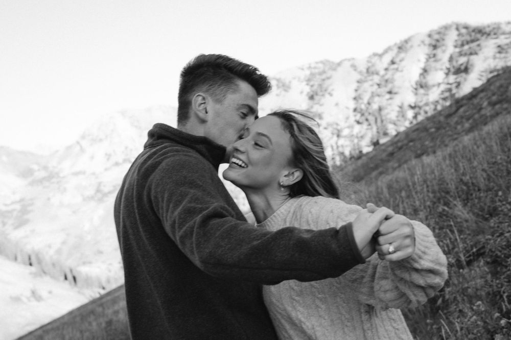 Black and white photo of couple embracing and laughing in snowy mountain setting.