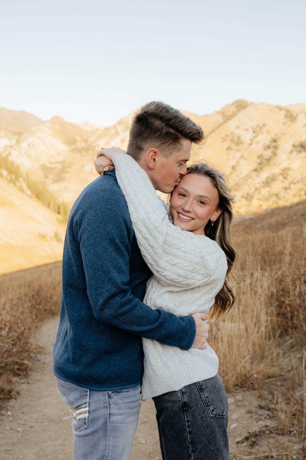 Man kissing woman's temple while embracing on a scenic mountain trail at sunset.