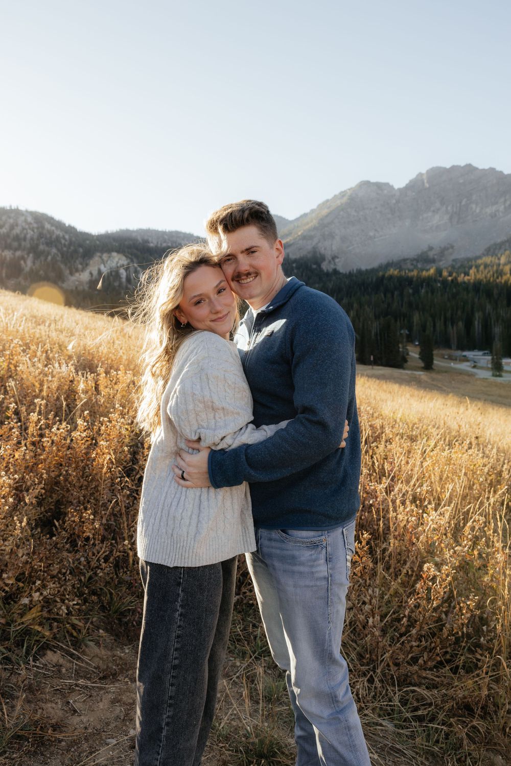 A couple embraces in a golden field with mountain backdrop during a fall engagement photo session.