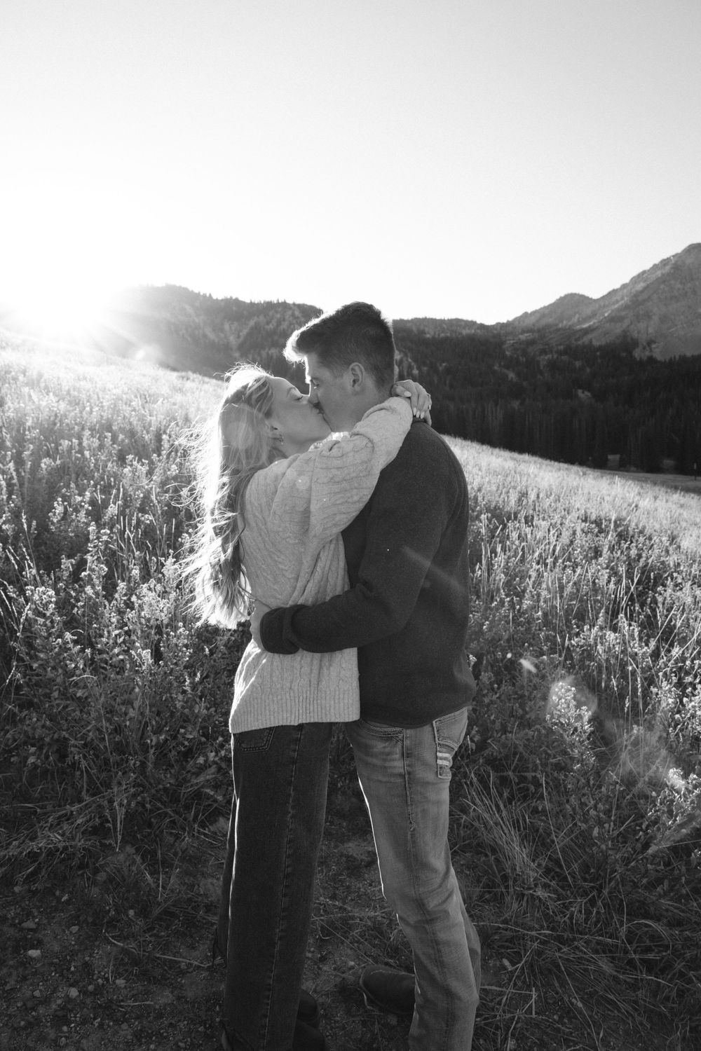 Black and white photo of couple kissing in sunlit mountain meadow at sunset.