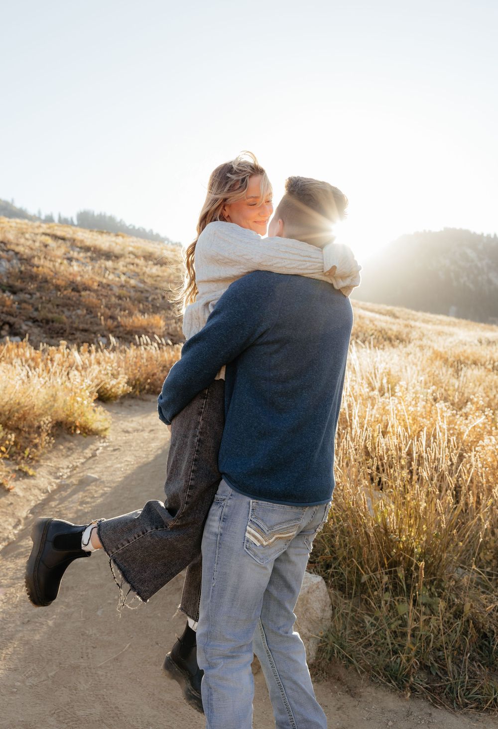 A man lifts a woman in a sunlit golden field during an outdoor couples engagement photo session.