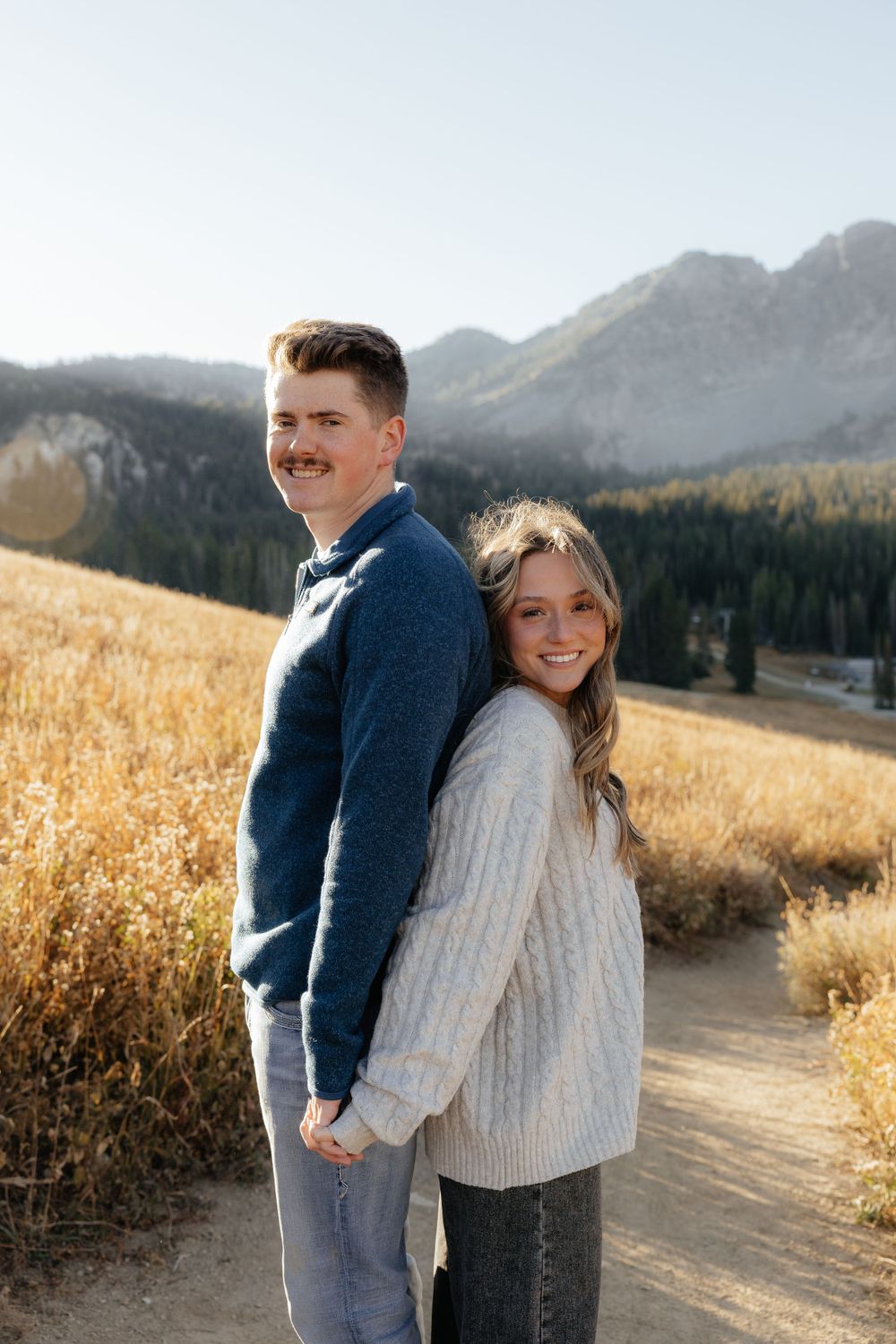 A smiling couple poses back-to-back on a mountain trail surrounded by autumn golden grass.