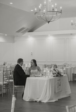 Black and white photo of bride and groom seated at sweetheart table in elegant wedding reception hall with chandelier.