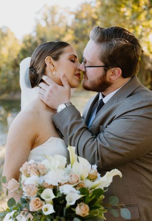 A romantic wedding couple shares a tender kiss while holding a bouquet of blush roses and white lilies in golden sunlight.