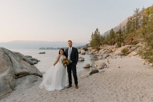 A couple in wedding attire stands on a rocky beach with mountains and lake in the background during golden hour.