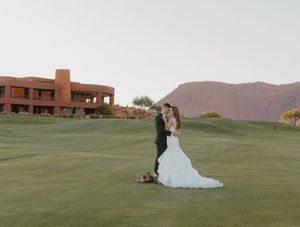 Two people embrace on a golf course with dramatic red rock mountains and a brick building in the background.