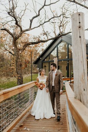 A couple stands on a wooden deck walkway outside a modern A-frame cabin surrounded by autumn trees.