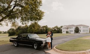 A classic black vintage car is parked on a curved driveway in front of a grand white mansion.