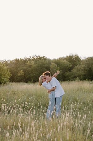 A couple embraces tenderly in a sun-lit meadow filled with tall grass and wildflowers at golden hour.