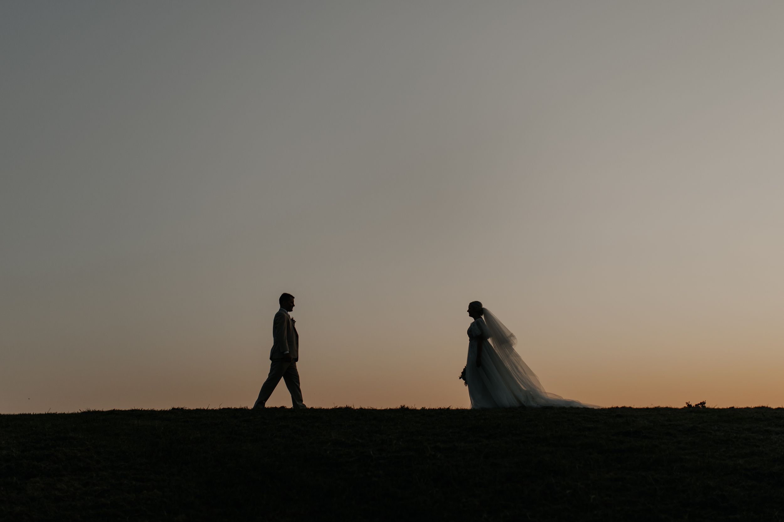 Silhouettes of bride and groom walking apart on hilltop against dusky sky at sunset.