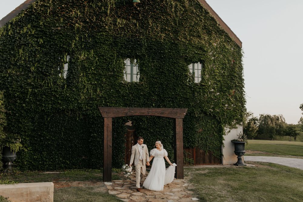 A couple exits a rustic brick building covered in lush green ivy on a sunny day.