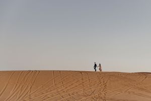 Two silhouetted figures stand atop a desert sand dune against a hazy sky during golden hour.