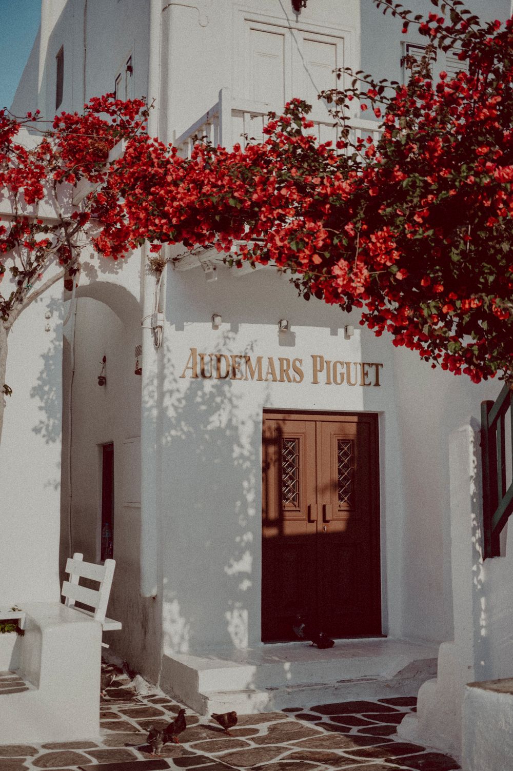 White Mediterranean building with Audemars Piguet signage framed by vibrant red bougainvillea flowers.