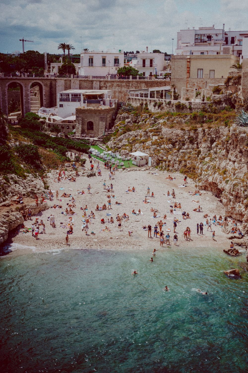 Crowded beach cove surrounded by white buildings and rocky cliffs in a Mediterranean coastal town.