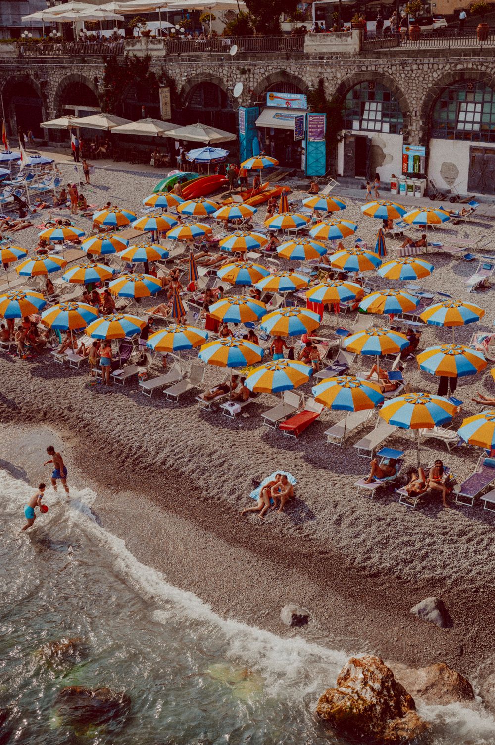 Beach scene with rows of orange and blue striped umbrellas along the shoreline near historic stone arches.