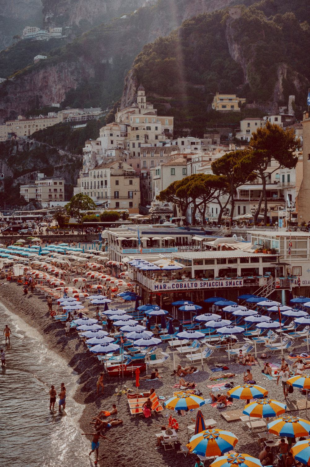Picturesque view of Amalfi Coast with white buildings cascading down cliffs and colorful beach umbrellas below.