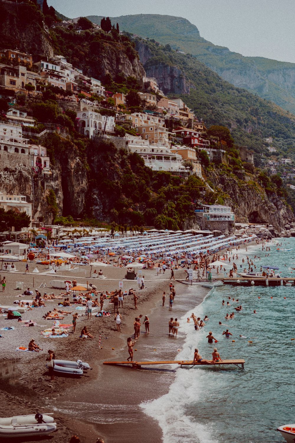 Beach scene at base of steep cliffs with swimmers, boats and beach umbrellas dotting the shoreline.