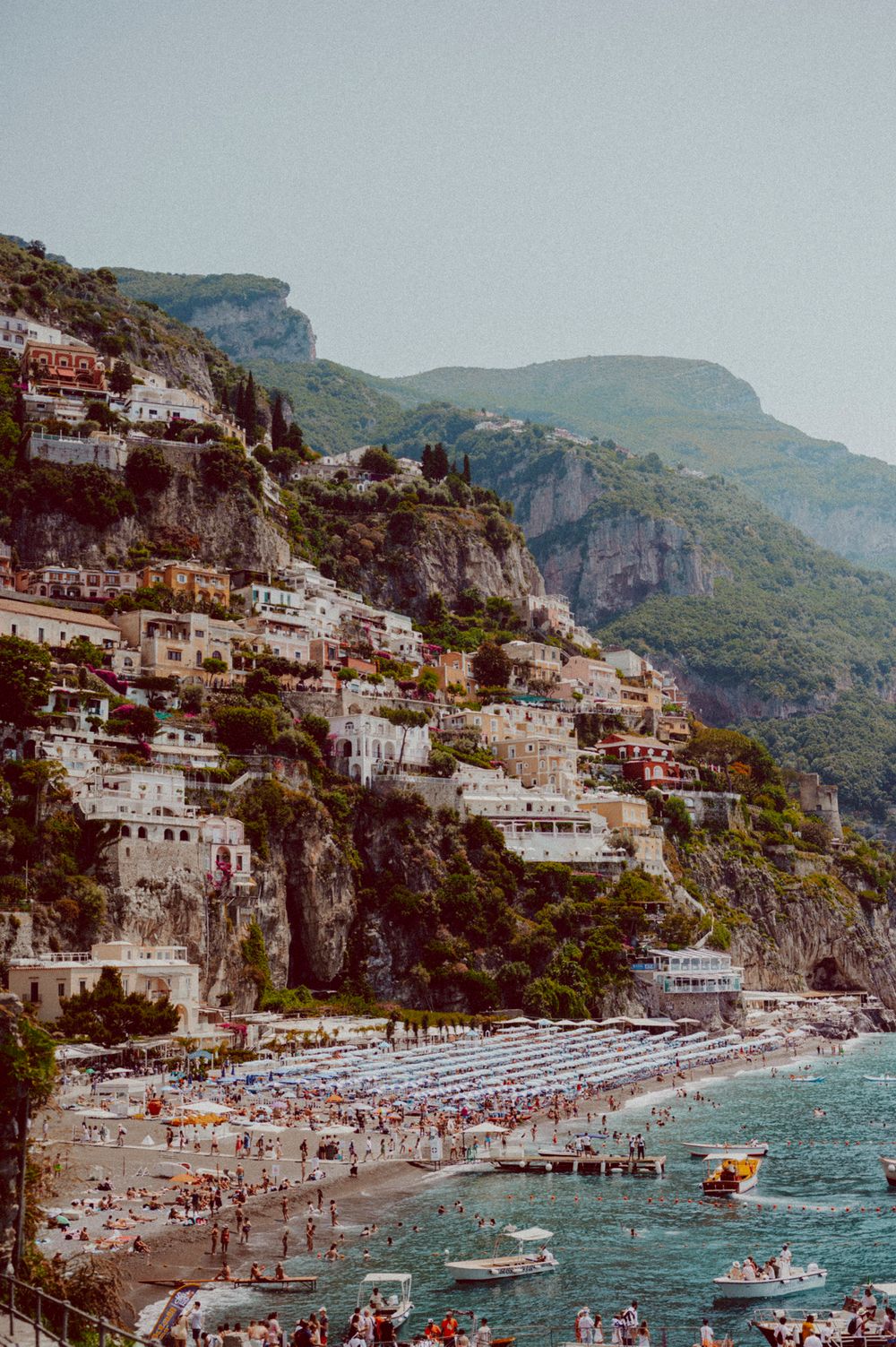 Panoramic view of Positano's hillside architecture and crowded beach along turquoise Mediterranean waters.