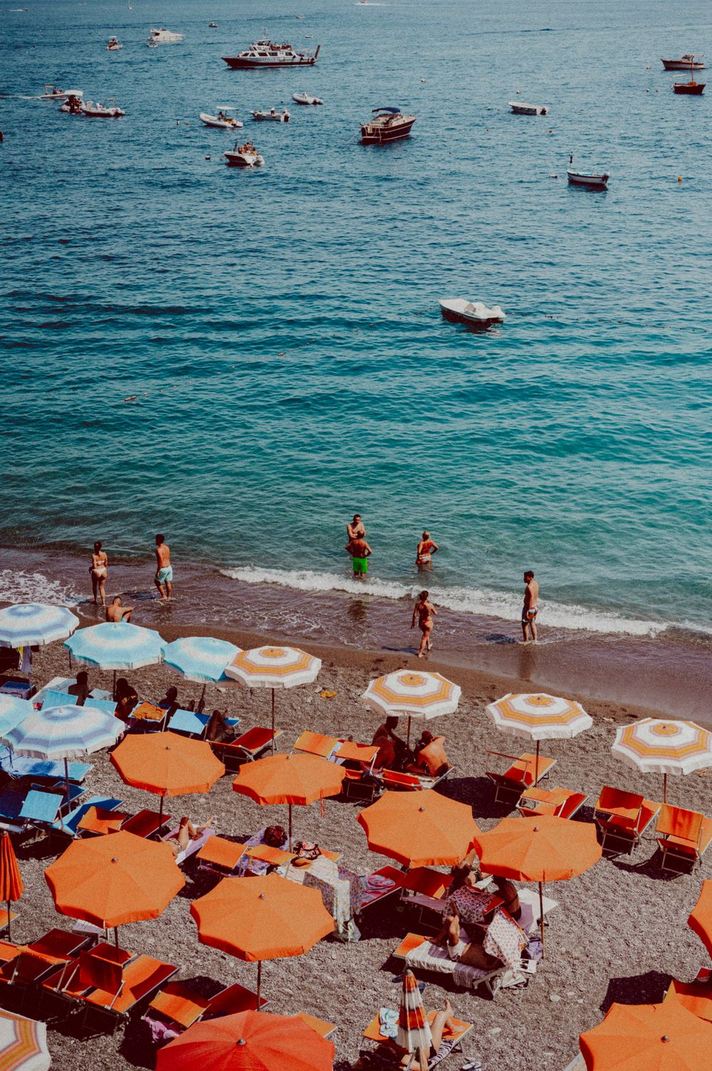 Beach scene with vibrant orange and white umbrellas lining the shore as people swim in turquoise waters.