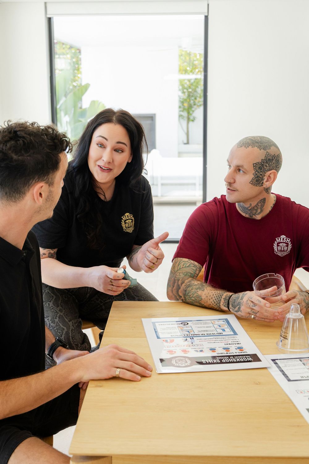 Three people sit around a wooden table having a business discussion with documents in a bright office setting.