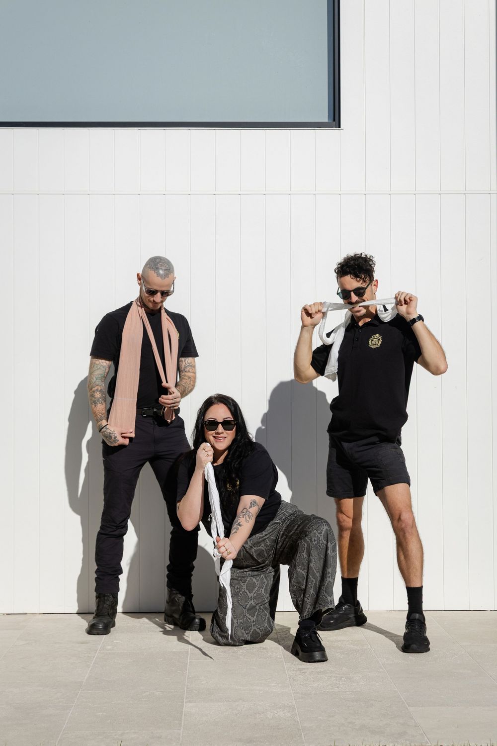 Three people in black outfits pose against a white wall while holding compression bandages. Mesh Education fun branding photography