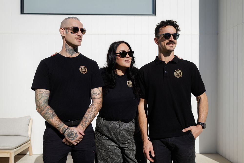 Group wearing matching black outfits pose together against white walls in modern minimal setting.