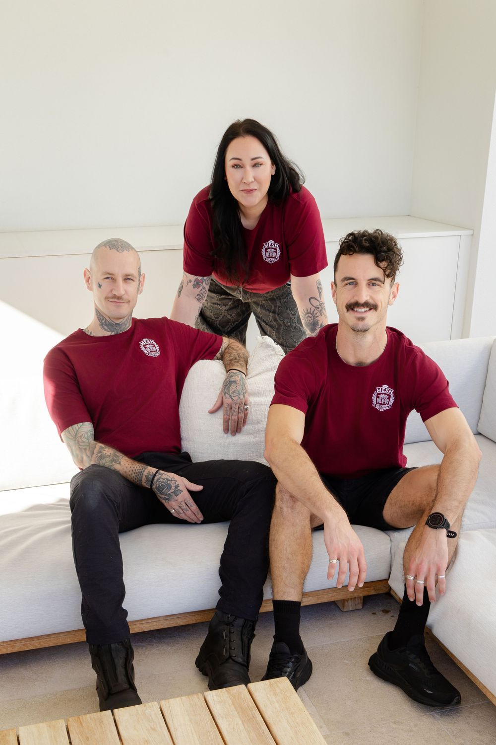 Three team members in matching burgundy polo shirts pose together on a white sofa.
