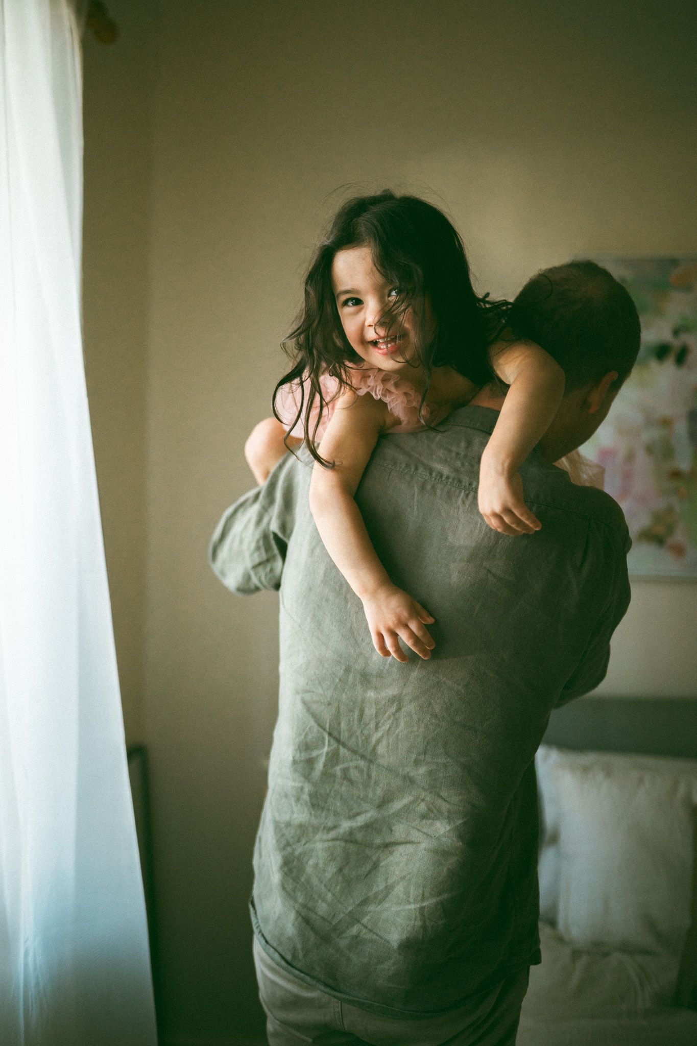 A parent in an off-shoulder brown dress holds a sleeping baby wrapped in a white blanket near a staircase.