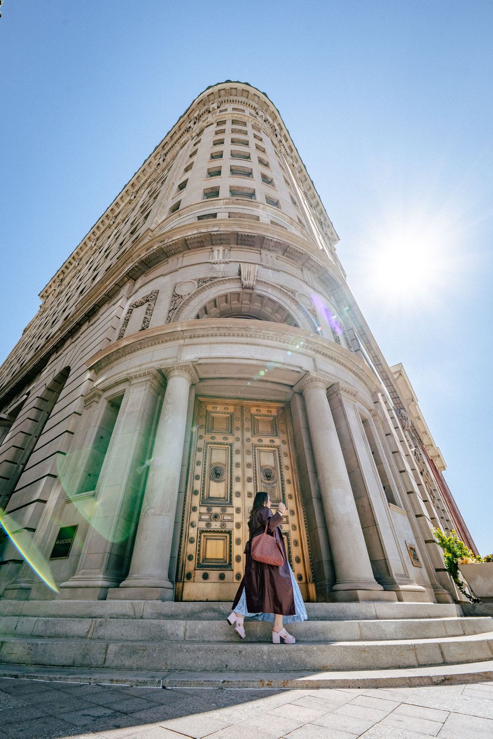 A person stands at impressive entrance of historic Flatiron-style building with ornate architecture and tall curved facade.