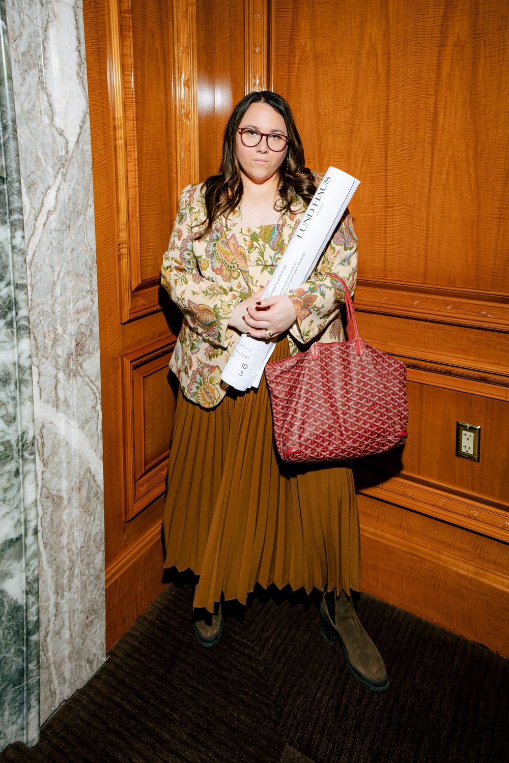 A person in a floral blazer and brown pleated skirt holds architectural plans while standing by wood-paneled walls.