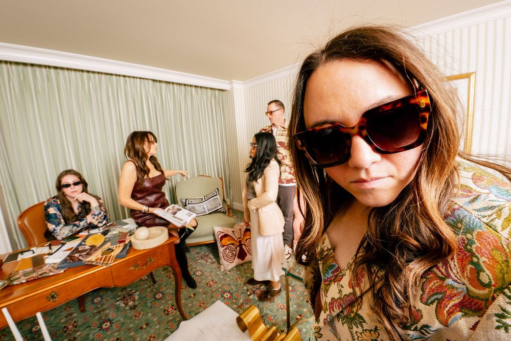 Woman in sunglasses poses with large collection of colorful shoes displayed on wooden furniture.