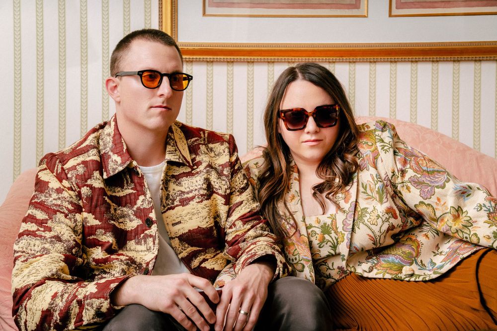 Two people wearing sunglasses and floral patterned clothing sit together on a retro upholstered sofa.