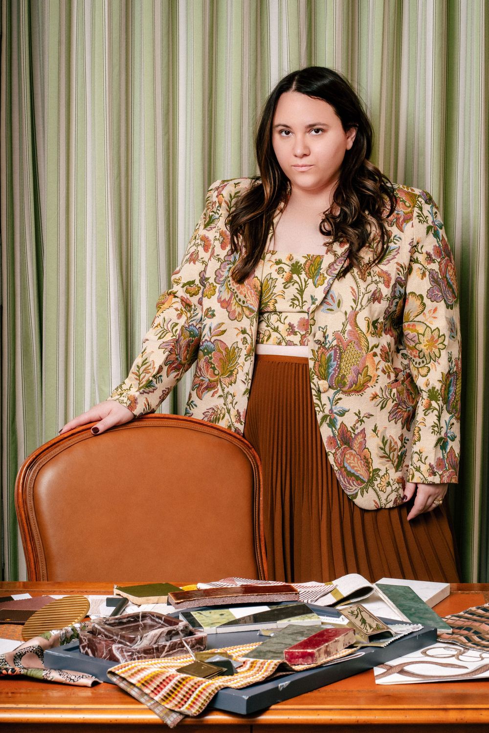 Interior photo of person in floral blazer and brown skirt standing by leather chair with fabric samples on wooden table.