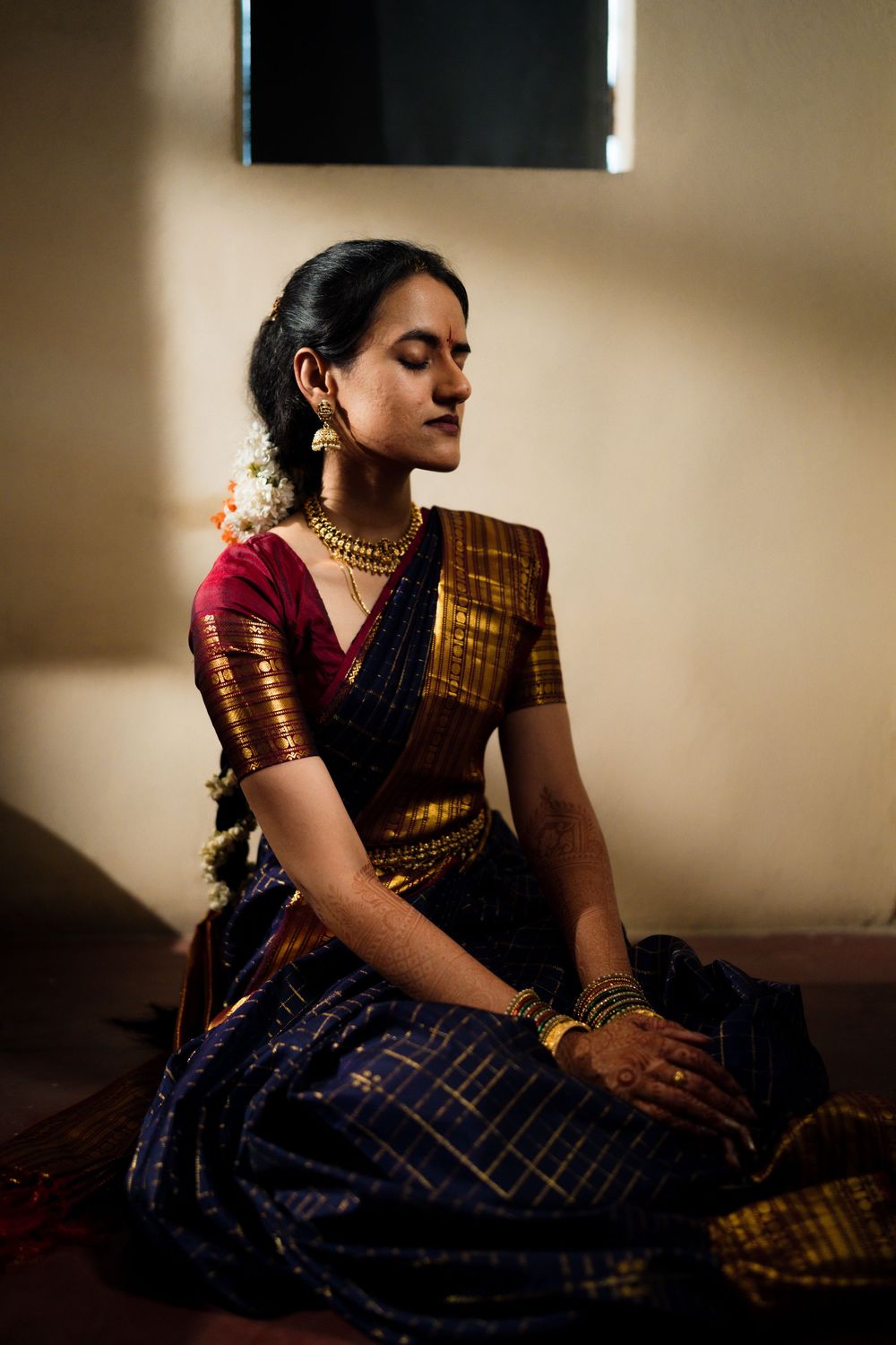 Person in traditional Indian attire with gold jewelry sits peacefully in meditation pose wearing a blue and maroon silk saree.