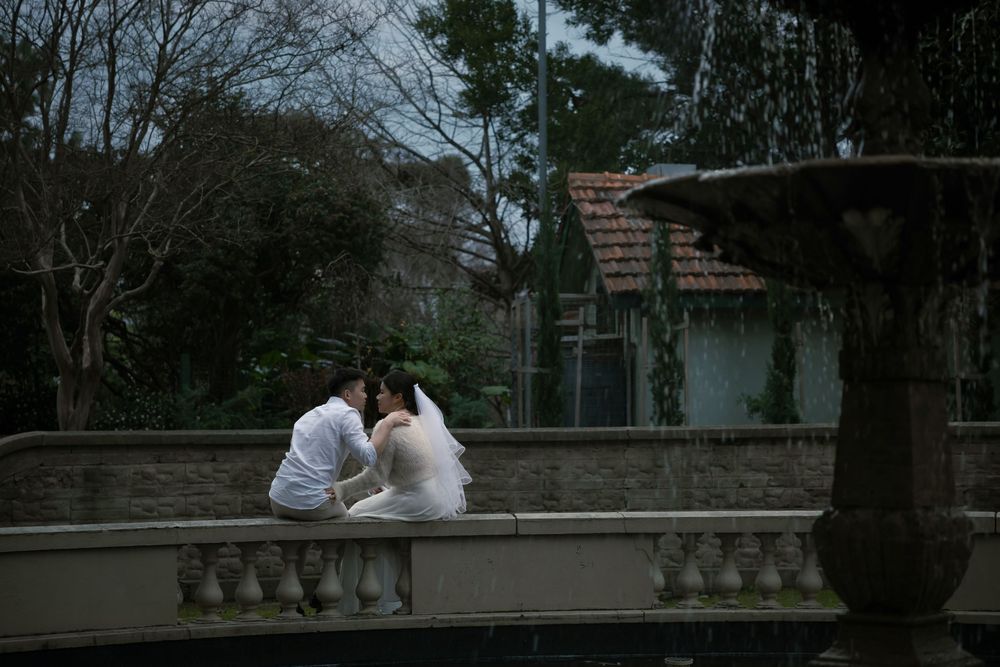 A romantic couple in white attire sits close together on a stone balustrade at dusk surrounded by trees.