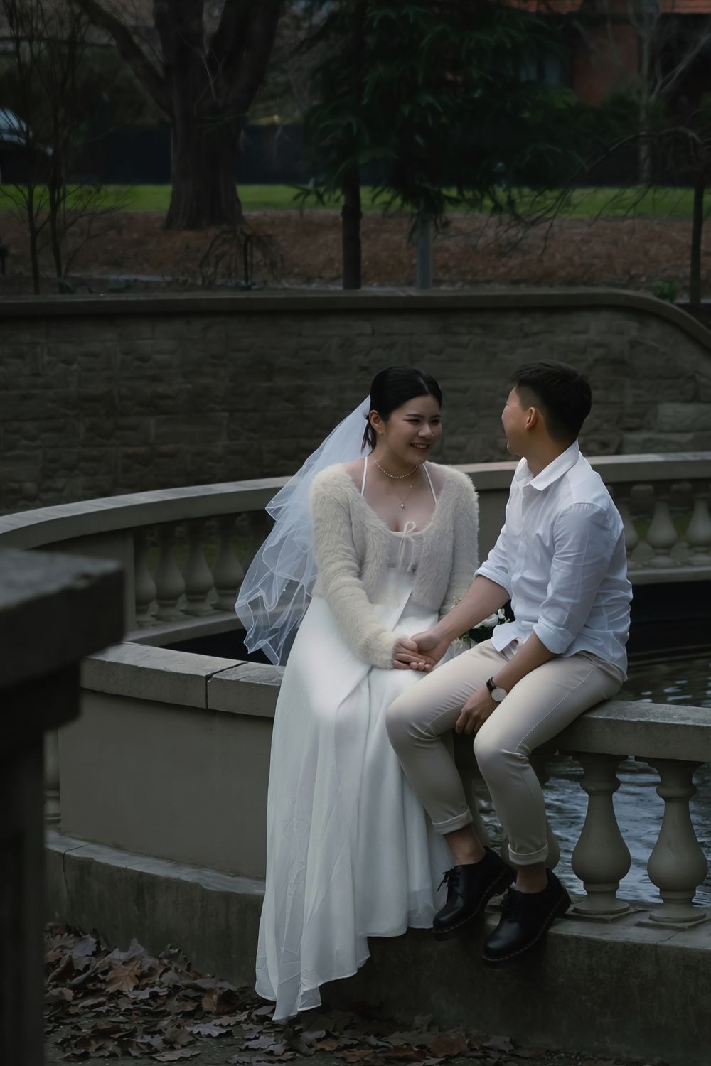 A couple in wedding attire sits intimately on stone steps in a park during an overcast day.