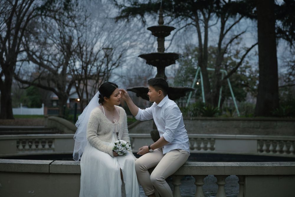 Two people in wedding attire share an intimate moment on stone benches in an outdoor garden setting.