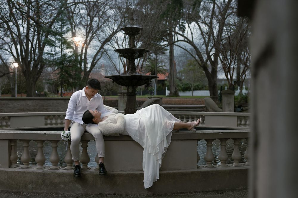A romantic couple in wedding attire poses beside an ornate fountain in a park at dusk.