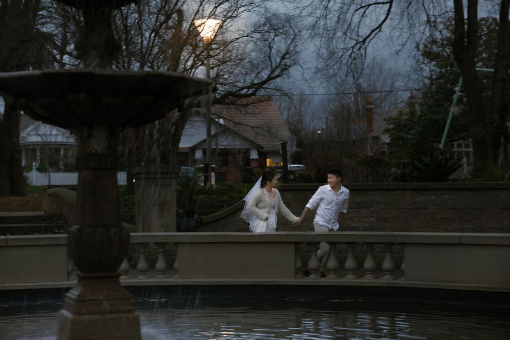 Two people in white clothing hold hands while walking along a stone balustrade near a fountain at dusk.