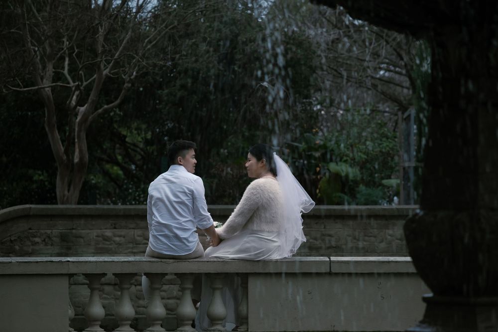 A couple in wedding attire sits together on a stone balustrade in a tranquil garden setting.