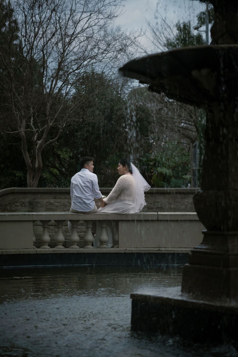 Two people sit together on a stone wall overlooking a tranquil pond at twilight.