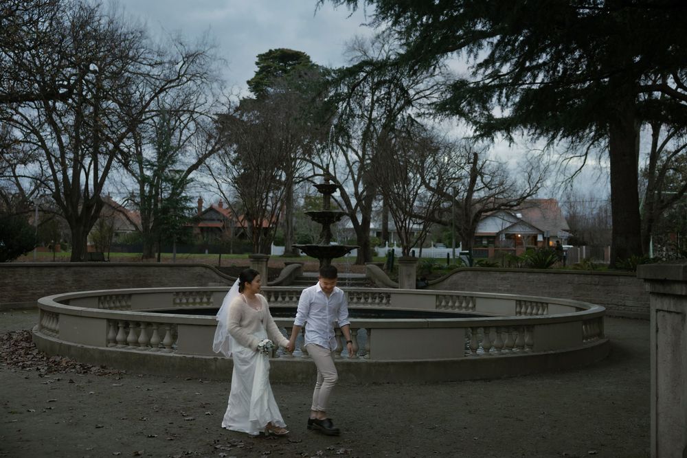 A couple stands on a curved stone terrace overlooking a park at dusk with bare winter trees in the background.