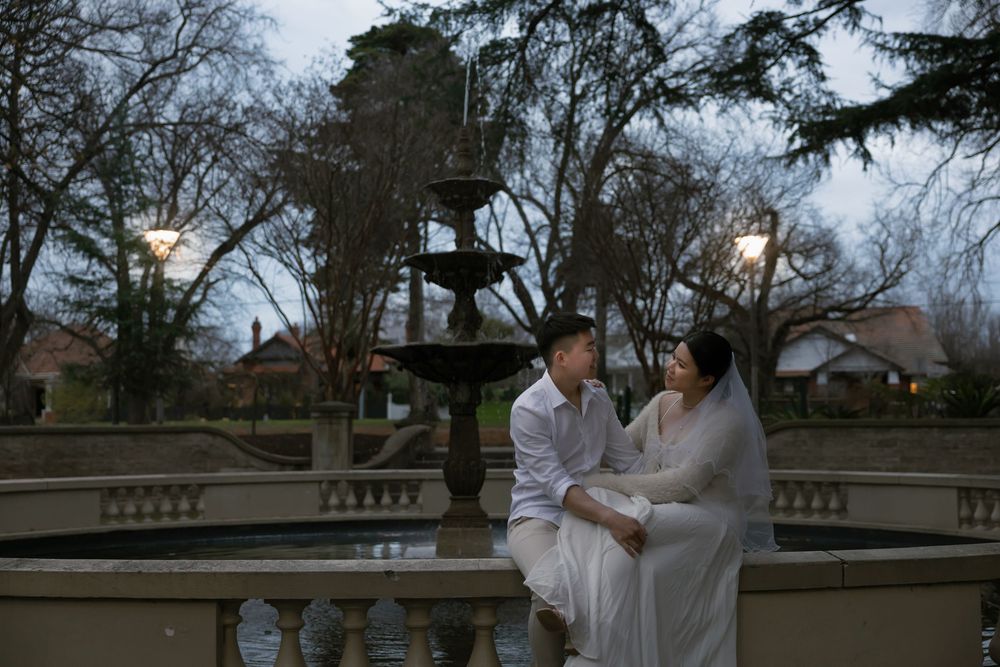 Two people in white outfits share an intimate moment while sitting on an ornate stone railing in a garden setting.