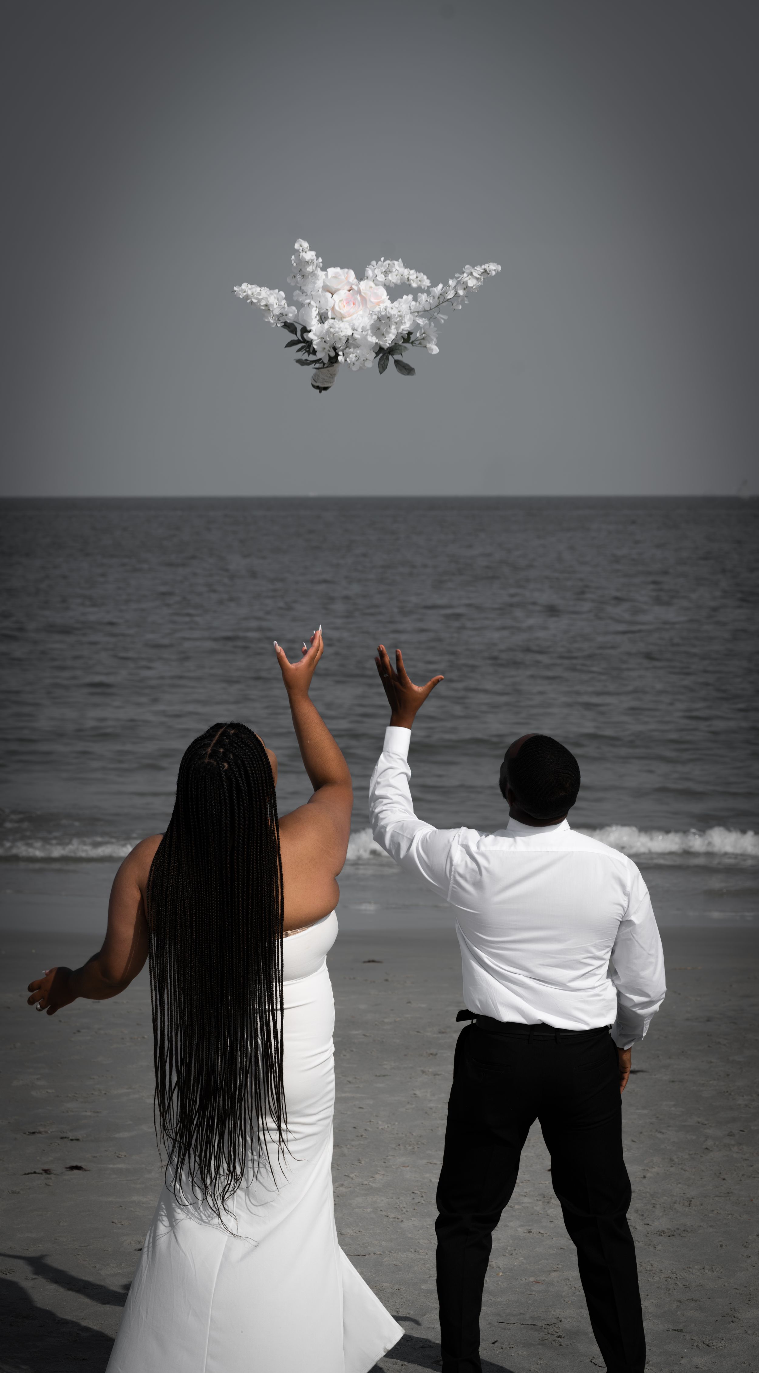 Two people in white and black attire throw a bouquet of flowers at the beach against a moody gray sky.