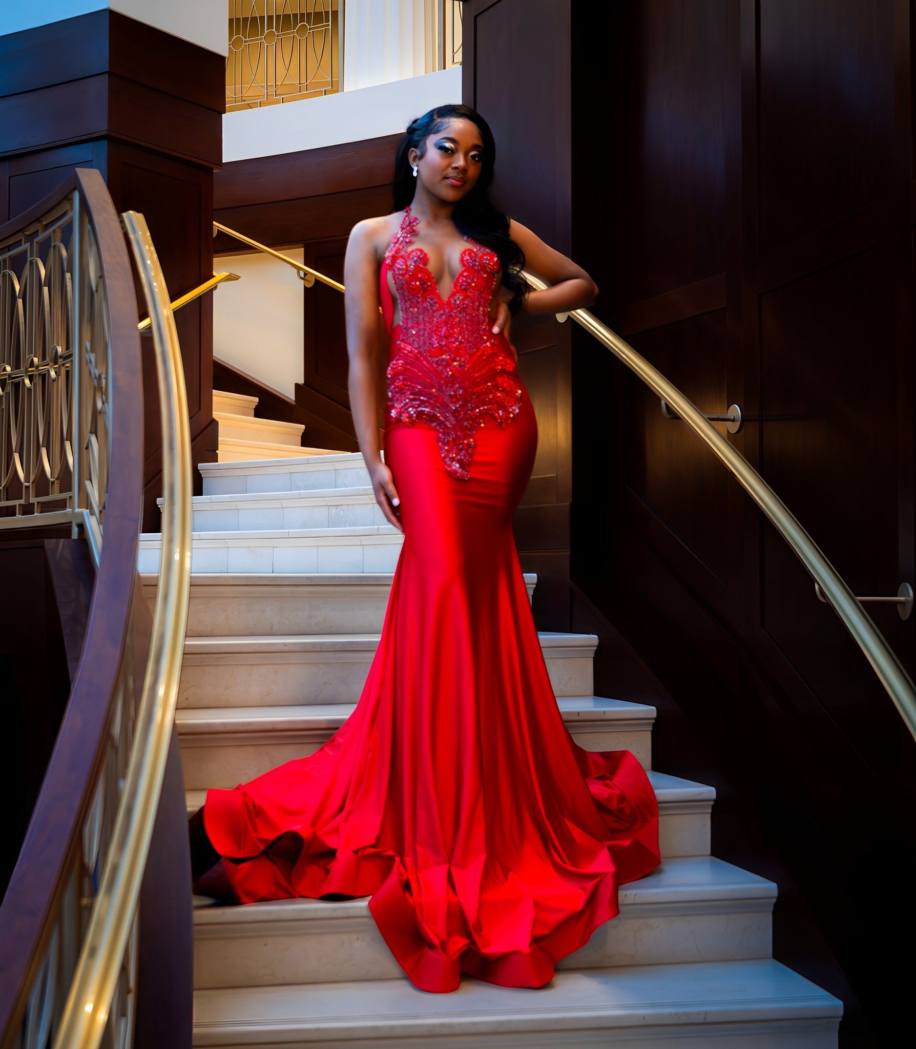 Someone wearing a stunning red mermaid gown with crystal details poses on an elegant staircase with gold railings.