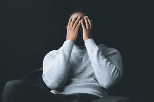 A figure in a gray sweatshirt sits with hands covering their face in a dark setting.