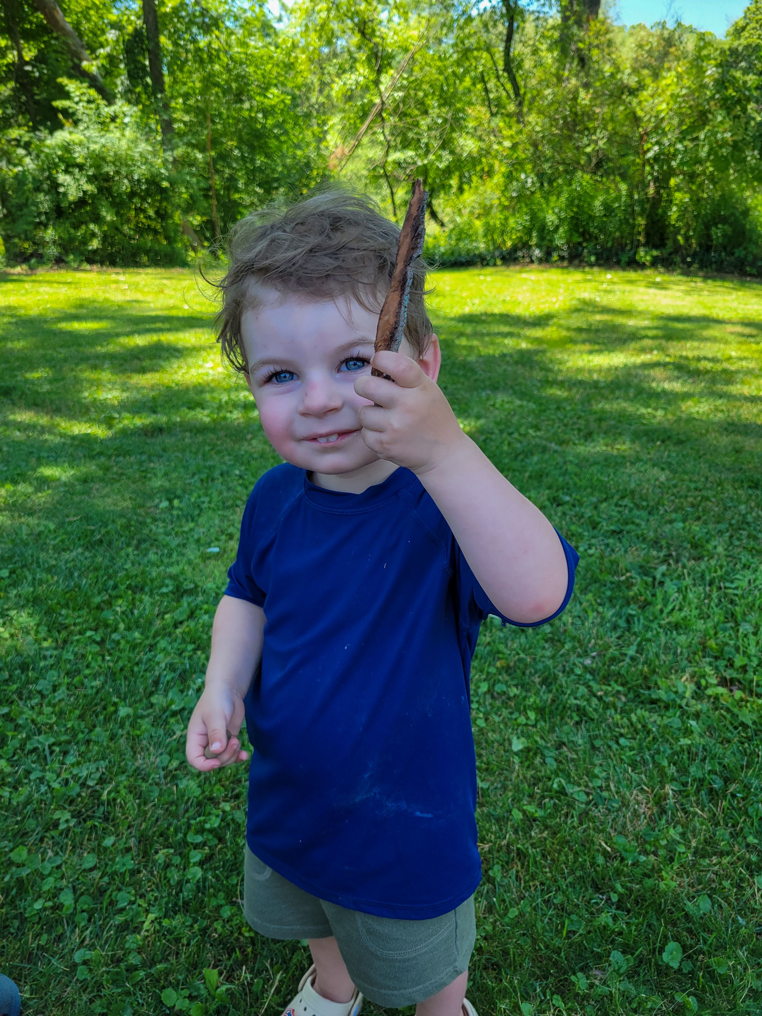 Child plays in a grassy backyard wearing a blue shirt on a bright summer day.