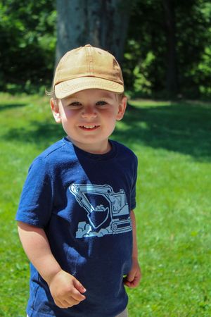 Young child wearing a navy t-shirt with construction vehicle design and tan cap playing outdoors on grass.